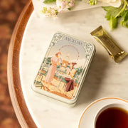 Angelina dark chocolate crispy crepes tin placed on a marble table beside a cup of tea and a wrapped chocolate, creating an elegant and indulgent tea-time scene.