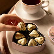 A hand picking a gold-wrapped heart-shaped chocolate from an open Angelina heart tin beside a cup of tea.