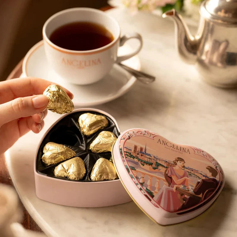 A person holding a gold-wrapped heart chocolate next to an open Angelina heart tin and a cup of tea.