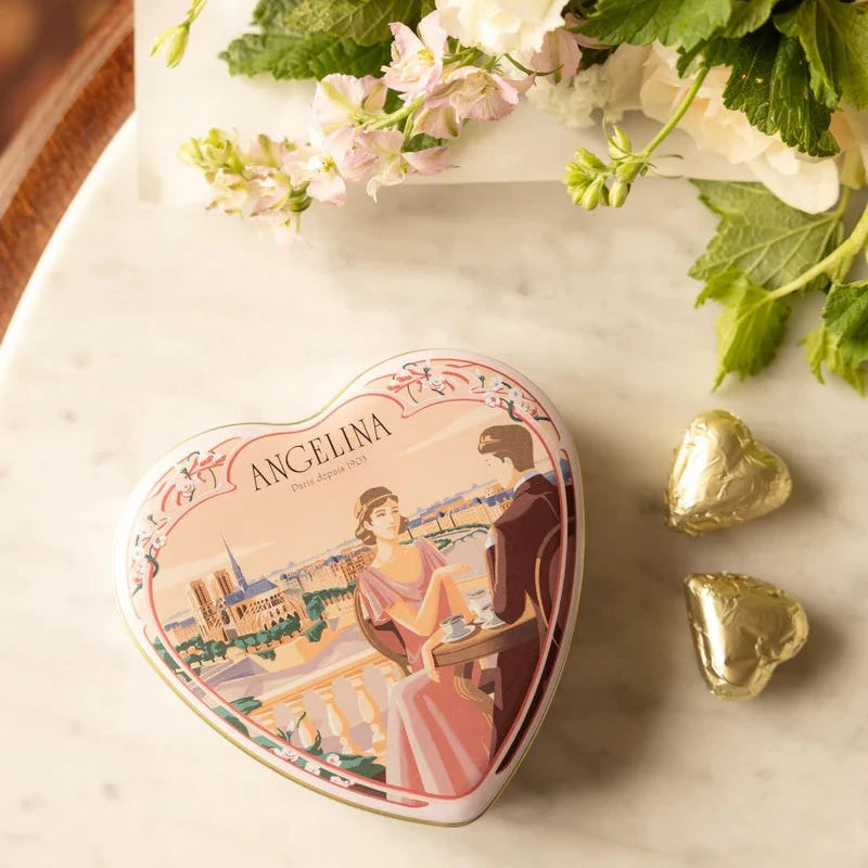 A closed Angelina heart tin placed on a marble table with flowers and two gold-wrapped chocolates beside it.