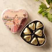 Open Angelina heart tin filled with gold-wrapped chocolates shown beside its illustrated lid on a marble surface.