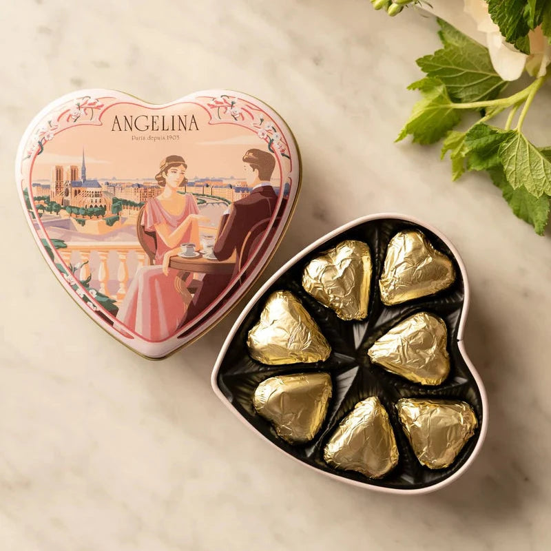 Open Angelina heart tin filled with gold-wrapped chocolates shown beside its illustrated lid on a marble surface.