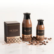 A luxurious Angelina Paris hot chocolate collection displayed on a marble surface, including the iconic black and gold metal tin of hot chocolate mix and two glass bottles of ready-to-drink rich chocolate beverage, surrounded by cocoa beans.
