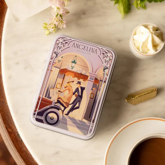 A tin of Angelina Paris Mini Giandujas placed on a marble table beside a cup of hot chocolate, whipped cream, and mini gianduja.