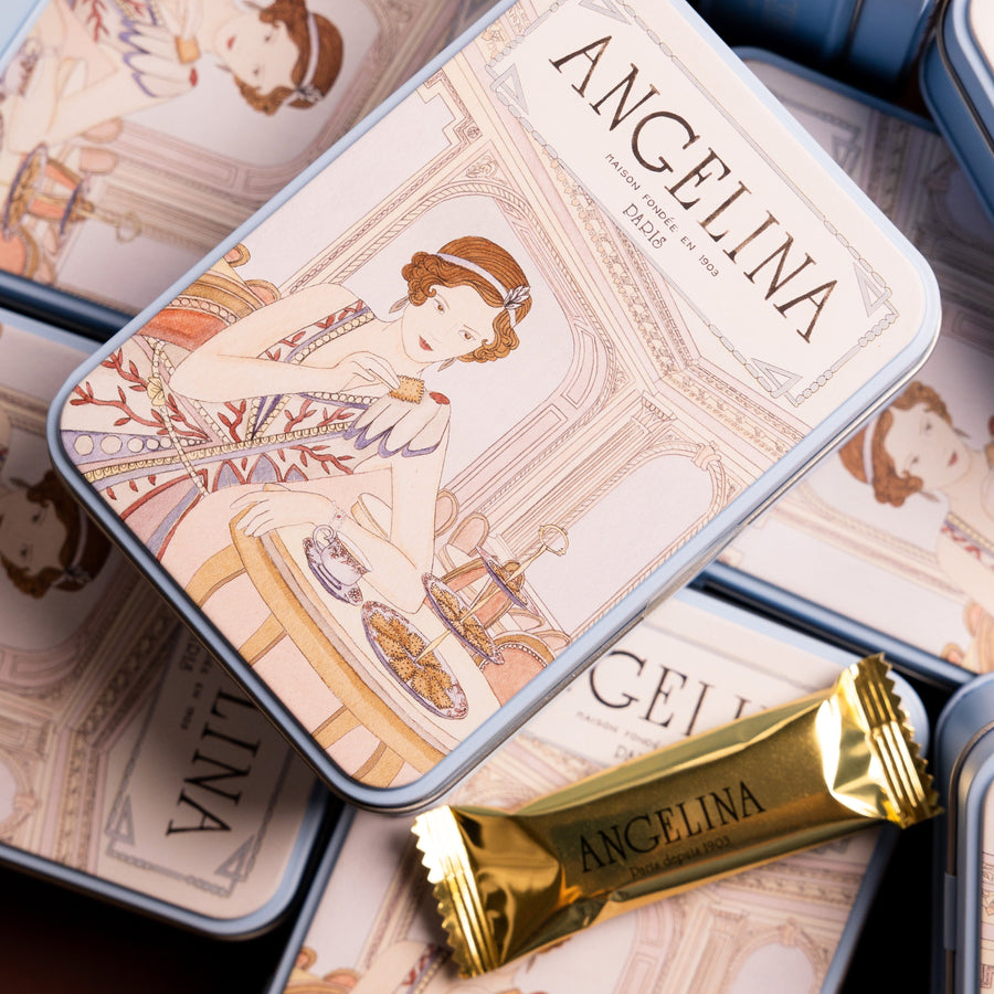 Stacked Angelina tins featuring an illustrated Parisian tea salon scene, with a wrapped dark chocolate crispy crepe in front.