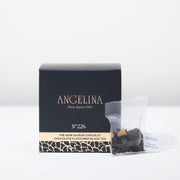 A black square box of Angelina Paris tea sits on a white surface beside a clear tea sachet containing chocolate flavored black tea leaves. The packaging, labeled “Chocolate Flavoured Black Tea – No. 226,” features gold Art Deco patterns and minimalist typography, conveying a luxurious and sophisticated Parisian style.