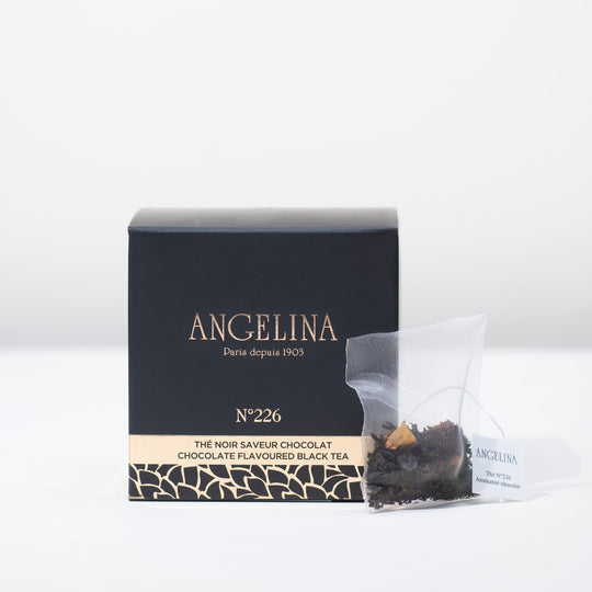A black square box of Angelina Paris tea sits on a white surface beside a clear tea sachet containing chocolate flavored black tea leaves. The packaging, labeled “Chocolate Flavoured Black Tea – No. 226,” features gold Art Deco patterns and minimalist typography, conveying a luxurious and sophisticated Parisian style.