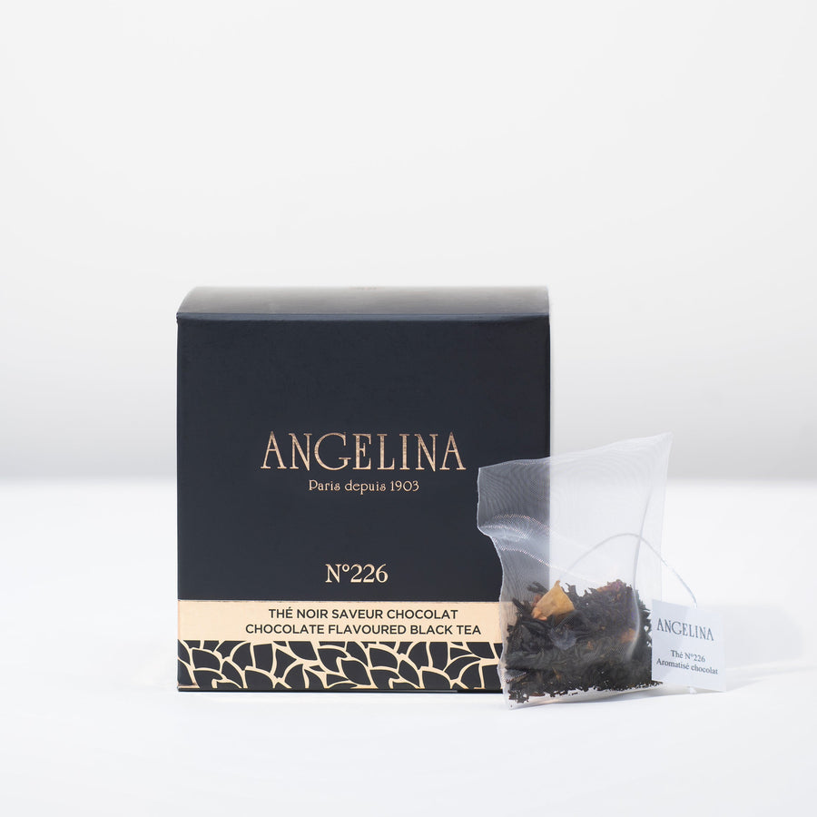 A black square box of Angelina Paris tea sits on a white surface beside a clear tea sachet containing chocolate flavored black tea leaves. The packaging, labeled “Chocolate Flavoured Black Tea – No. 226,” features gold Art Deco patterns and minimalist typography, conveying a luxurious and sophisticated Parisian style.