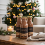 Elegant holiday lifestyle scene featuring three Angelina Paris hot chocolate bottles on a wooden table, with a softly lit Christmas tree and cozy neutral decor in the background.
