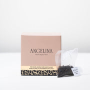 A soft pink square box of Angelina Paris tea is displayed on a white surface, accompanied by a transparent tea sachet filled with loose black tea leaves. The box reads “Mont Blanc Flavoured Black Tea” in English and French, with elegant gold and black Art Deco detailing at the base, expressing a refined Parisian aesthetic.