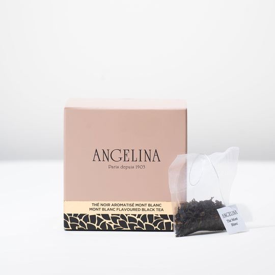 A soft pink square box of Angelina Paris tea is displayed on a white surface, accompanied by a transparent tea sachet filled with loose black tea leaves. The box reads “Mont Blanc Flavoured Black Tea” in English and French, with elegant gold and black Art Deco detailing at the base, expressing a refined Parisian aesthetic.