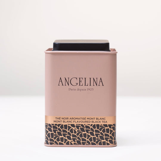A pale pink metal tea tin from Angelina Paris is displayed on a white surface. The tin features a minimalist Art Deco design with gold and black leaf motifs at the bottom and the text “Mont Blanc Flavoured Black Tea” printed in English and French. It conveys a refined and elegant Parisian aesthetic.