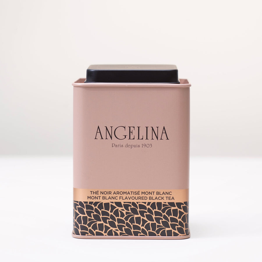 A pale pink metal tea tin from Angelina Paris is displayed on a white surface. The tin features a minimalist Art Deco design with gold and black leaf motifs at the bottom and the text “Mont Blanc Flavoured Black Tea” printed in English and French. It conveys a refined and elegant Parisian aesthetic.