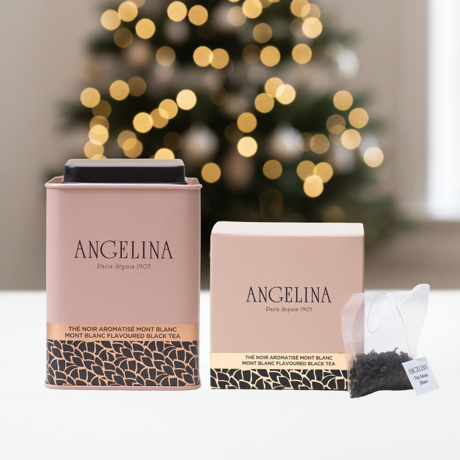 A refined holiday image featuring two Mont Blanc Flavored Black Tea products from Angelina Paris. On the left is a pale pink metal tea tin, and on the right, a matching square box with a transparent tea sachet placed in front. The background shows a softly blurred Christmas tree with warm golden lights, creating an elegant and festive Parisian atmosphere.