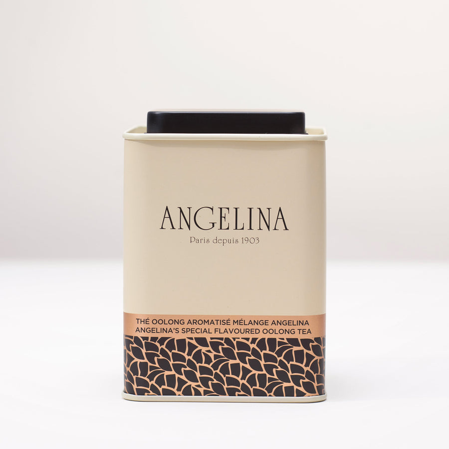 A cream-colored metal tea tin from Angelina Paris is shown on a white surface. The tin has a clean, elegant design with black and gold Art Deco patterns at the base and the label “Angelina’s Special Flavoured Oolong Tea” in English and French, reflecting the brand’s classic Parisian sophistication.