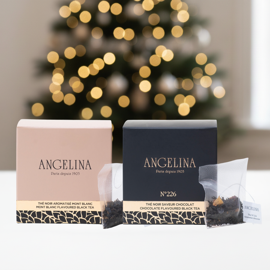 Two elegant tea bag boxes of Angelina Paris teas are displayed side by side on a white surface, with a softly blurred Christmas tree and warm festive lights in the background. The left box is a pale pink labeled “Mont Blanc Flavoured Black Tea,” and the right box is dark navy labeled “Chocolate Flavoured Black Tea – No. 226.” Each box is accompanied by a transparent tea sachet in front, evoking a refined, holiday gift atmosphere.