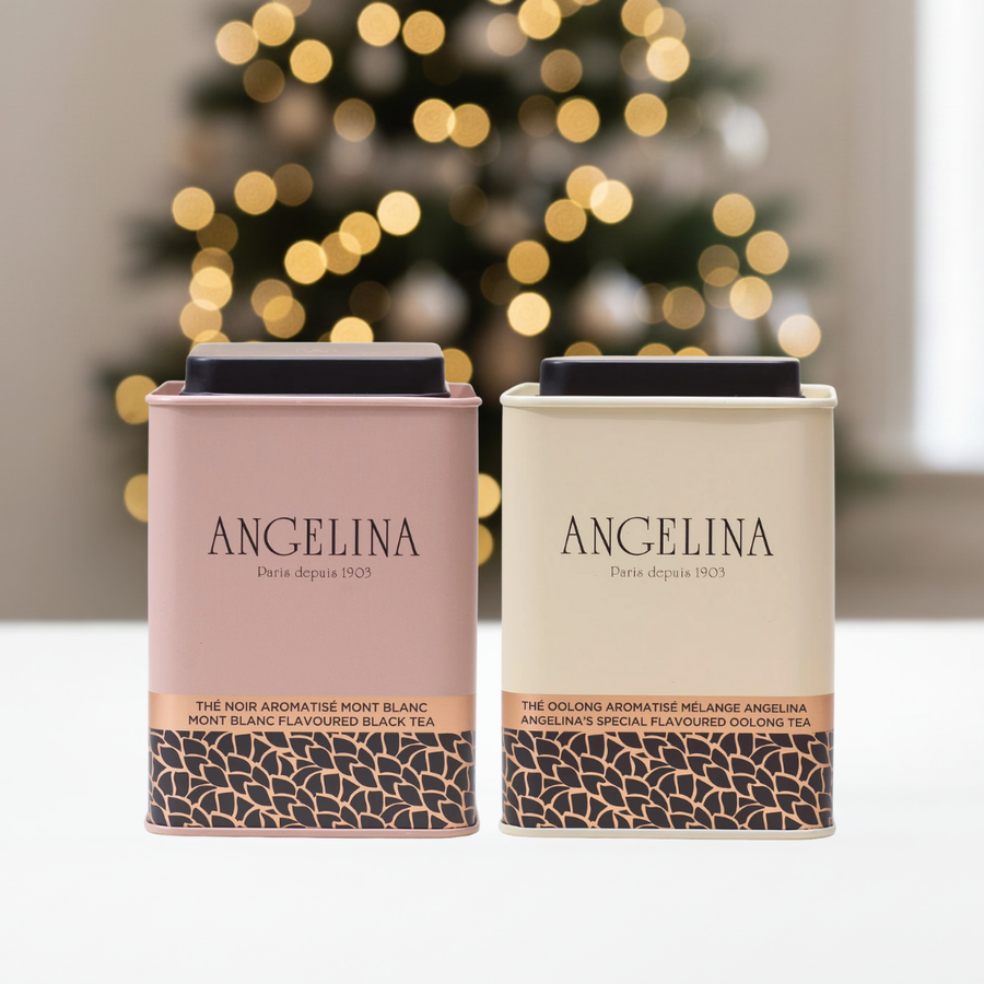 Two elegant metal tea tins from Angelina Paris are displayed on a white surface with a softly blurred Christmas tree and warm golden lights in the background. The left loose leaf tea tin is pale pink labeled “Mont Blanc Flavoured Black Tea,” and the right loose leaf tea tin is light cream labeled “Angelina’s Special Flavoured Oolong Tea.” Both feature refined gold and black Art Deco patterns at the base, evoking Parisian sophistication and a festive holiday mood.